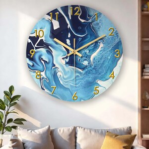 Op de afbeelding: Ronde wandklok met een blauw-wit abstract ontwerp. De wijzerplaat heeft goudkleurige cijfers en wijzers. De klok is aan een witte muur bevestigd, met een bank en een plant op de voorgrond.