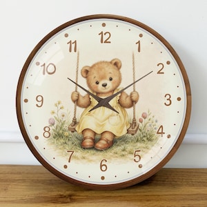 May include: A round wall clock with a dark wood frame and a cream-colored face. The clock features brown numerals from 1 to 12 and dark brown hands. The center illustration depicts a brown teddy bear in a yellow dress sitting on a rope swing.