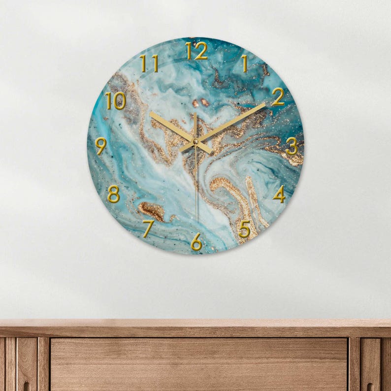 May include: Round wall clock with a marbled blue and gold design. The clock face features gold numerals and hands. The clock is mounted on a white wall above a wooden surface.