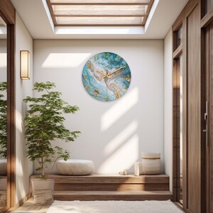 May include: A round wall clock with a blue and gold abstract design. The clock face features gold numerals and hands. The clock is mounted on a white wall in a well-lit room with a plant and wooden accents.