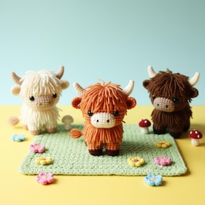 May include: Three crocheted Highland cow figures in cream, orange, and brown, with small horns and shaggy fur. They are on a green crocheted mat with flower and mushroom decorations, set against a yellow and blue background.