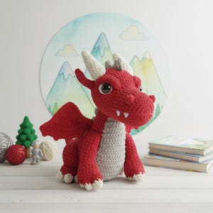May include: A handmade red and gray crochet dragon with white horns, wings, and claws. The dragon has large, round eyes and is sitting on a white surface. A watercolor mountain scene is in the background.