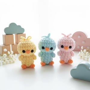 Könnte beinhalten: Drei gehäkelte Küken-Spielzeuge in Pastellfarben: Gelb, Blau und Rosa. Jedes Küken hat schwarze Augen, einen orangefarbenen Schnabel und orangefarbene Füße. Die Spielzeuge sind handgefertigt und stehen aufrecht.