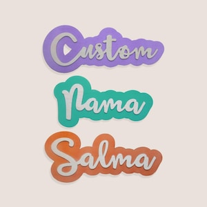 Op de afbeelding: Drie aangepaste naamborden in een gelaagd ontwerp. Het bovenste bord leest "Custom" in een zilveren scriptlettertype, tegen een paarse wolkachtige achtergrond. Het middelste bord leest "Nama" in een zilveren scriptlettertype, tegen een turquoise wolkachtige achtergrond. Het onderste bord leest "Salma" in een zilveren scriptlettertype, tegen een oranje wolkachtige achtergrond.
