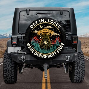 May include: Black Jeep with a spare tire cover featuring a UFO design. The cover has the text "GET IN, LOSER WE'RE DOING BUTT STUFF" around a graphic of a UFO beam abducting a person. The background includes mountains and trees.
