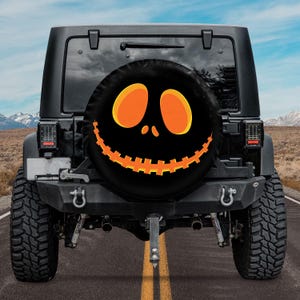 Puede incluir: Funda de rueda de repuesto negra con un diseño de cara de calabaza de Halloween, con ojos naranjas y boca dentada. La funda está montada en un Jeep negro, estacionado en una carretera con montañas al fondo. Un accesorio temático de Halloween.