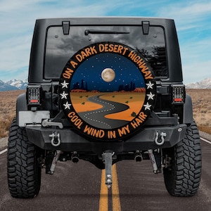 May include: Black Jeep with a spare tire cover featuring a desert highway scene under a full moon. The cover has the text "ON A DARK DESERT HIGHWAY COOL WIND IN MY HAIR" in a circular design.