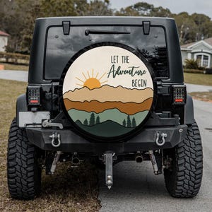 May include: A black Jeep with a spare tire cover featuring a mountain and sun design. The cover has the text "LET THE Adventure BEGIN" in green and white. The design includes shades of brown, green, and yellow.