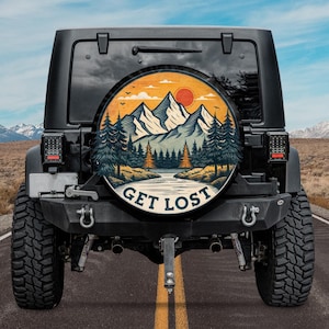 May include: Black Jeep with a spare tire cover featuring a mountain landscape design. The cover has a circular design with a mountain range, trees, and the text "GET LOST" in white. The background shows a road and a mountain range.
