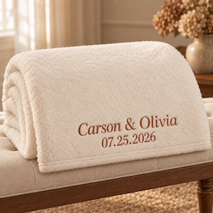 May include: A cream-colored, textured fleece blanket featuring the embroidered brown text Carson & Olivia and the date 07.25.2026. The blanket is folded neatly, showcasing its soft, plush material and the personalized lettering on the front.