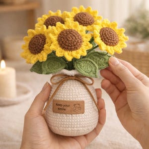May include: A crocheted bouquet of sunflowers in a white vase. The sunflowers have yellow petals and brown centers. The vase has a small tag that says "Keep your smile" with a bear illustration. The vase is tied with a brown string.