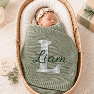 May include: A sage green knitted baby blanket featuring a large, grey and white gingham letter L with the name Liam embroidered in dark green cursive script. The blanket is wrapped around a sleeping infant wearing a floral headband in a basket.