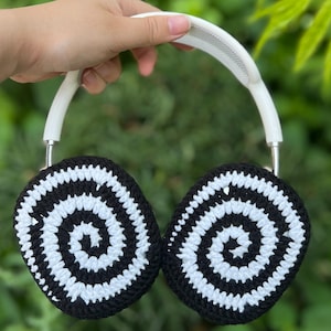 May include: A pair of headphones with black and white crocheted ear covers featuring a spiral design. The headband is white, and the headphones are held against a backdrop of green foliage.