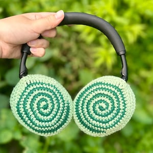 Könnte beinhalten: Gehäkelter Kopfhörer mit grünen und hellgrünen Spiralohrmuscheln und einem schwarzen Bügel. Die Ohrmuscheln sind rund und weisen ein detailliertes Spiralmuster auf. Der Kopfhörer wird vor einem verschwommenen grünen Hintergrund gehalten.