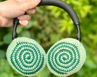 Könnte beinhalten: Gehäkelter Kopfhörer mit grünen und hellgrünen Spiralohrmuscheln und einem schwarzen Bügel. Die Ohrmuscheln sind rund und weisen ein detailliertes Spiralmuster auf. Der Kopfhörer wird vor einem verschwommenen grünen Hintergrund gehalten.
