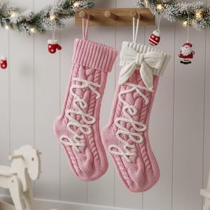 以下が含まれることがあります： ピンク色のケーブルニットのクリスマスストッキング2つ。白い筆記体で「Kelly」と刺繍されています。片方のストッキングには白いリボンが付いています。木製のラックに吊るされており、お祝いのオーナメントとガーランドで飾られています。