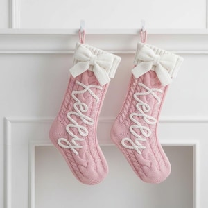 Può includere: Due calze di Natale rosa a trecce con fiocchi e polsini bianchi. Ogni calza ha il nome "Kelly" ricamato in corsivo bianco. Sono appese a ganci bianchi contro un muro bianco.