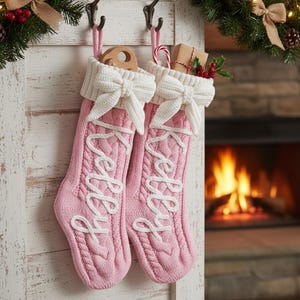Puede incluir: Dos calcetines navideños de punto rosa claro a juego colgados de ganchos de metal. Cada calcetín presenta un puño de punto trenzado blanco con un lazo blanco decorativo y el nombre Kelly bordado en letra cursiva blanca sobre el cuerpo rosa.