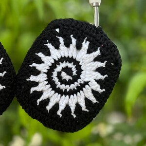 May include: Black and white crochet headphone covers. The covers feature a white spiral design on a black background. The crochet work is visible, and the covers are hanging against a blurred green background.