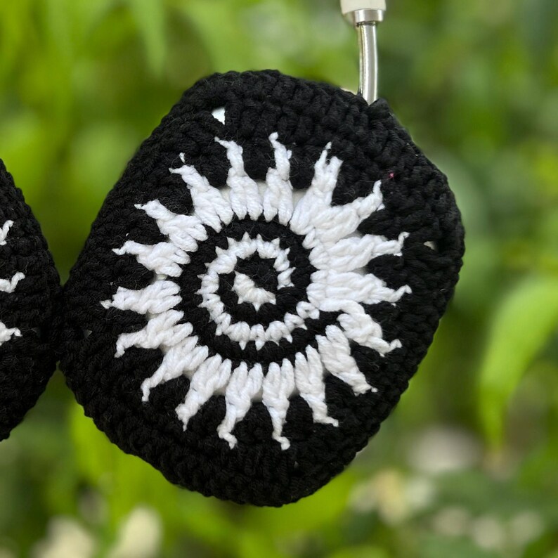 May include: Black and white crochet headphone covers. The covers feature a white spiral design on a black background. The crochet work is visible, and the covers are hanging against a blurred green background.