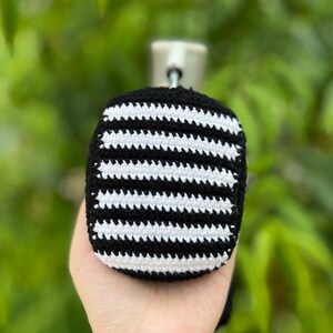 May include: A black and white striped crochet item, possibly a case or cover. The item is held in a hand, with a blurred green background of foliage. The stripes are horizontal and evenly spaced, creating a bold pattern.