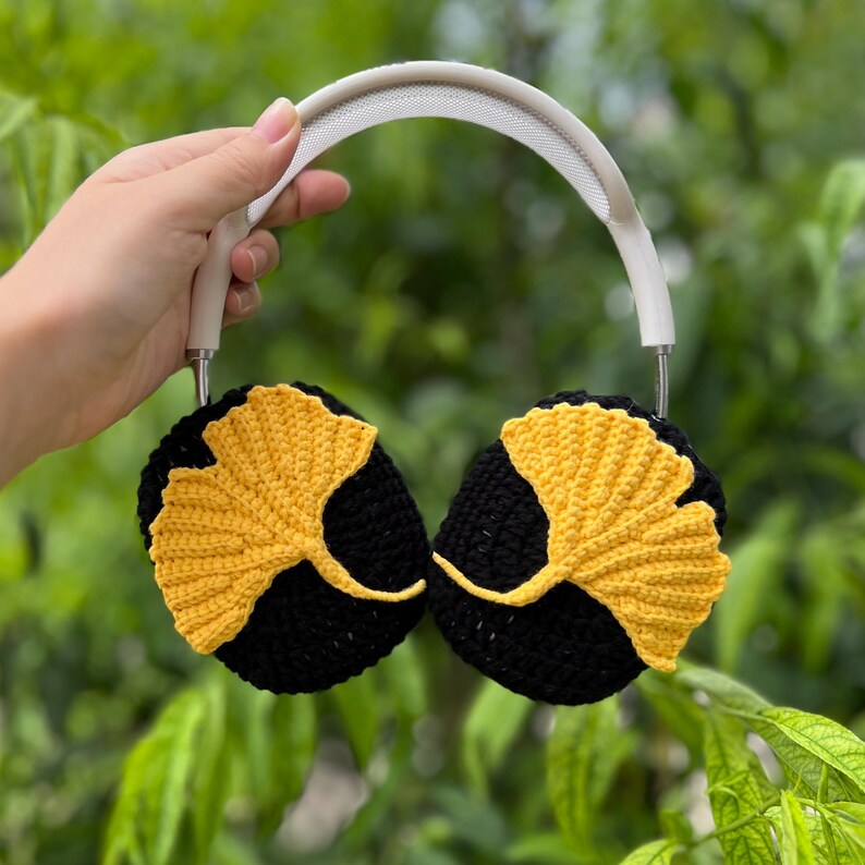 Pu&ograve; includere: Cuffie bianche e argentate con padiglioni neri, ciascuno ornato da un design a foglia di ginkgo gialla. Le foglie sono all'uncinetto, aggiungendo una trama unica. Lo sfondo &egrave; verde sfocato.