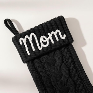 May include: Black knitted Christmas stocking with a cable knit design. The cuff features the word "Mom" in white script. Includes a loop for hanging. A festive holiday decoration.