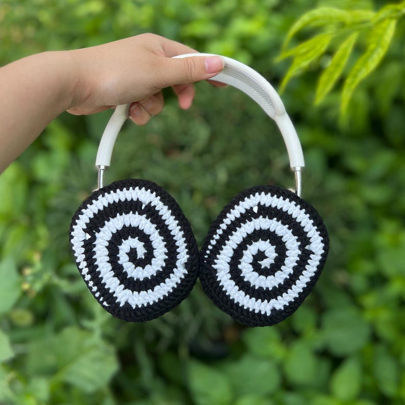 May include: A pair of headphones with black and white crocheted ear covers featuring a spiral design. The headband is white, and the headphones are held against a backdrop of green foliage.
