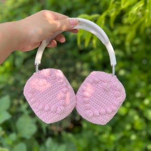 K&ouml;nnte beinhalten: Rosa geh&auml;kelte Kopfh&ouml;rerabdeckungen mit einem wei&szlig;en Stirnband. Die Ohrmuscheln haben ein strukturiertes Muster. Die Kopfh&ouml;rer werden vor einem gr&uuml;nen, belaubten Hintergrund gehalten.