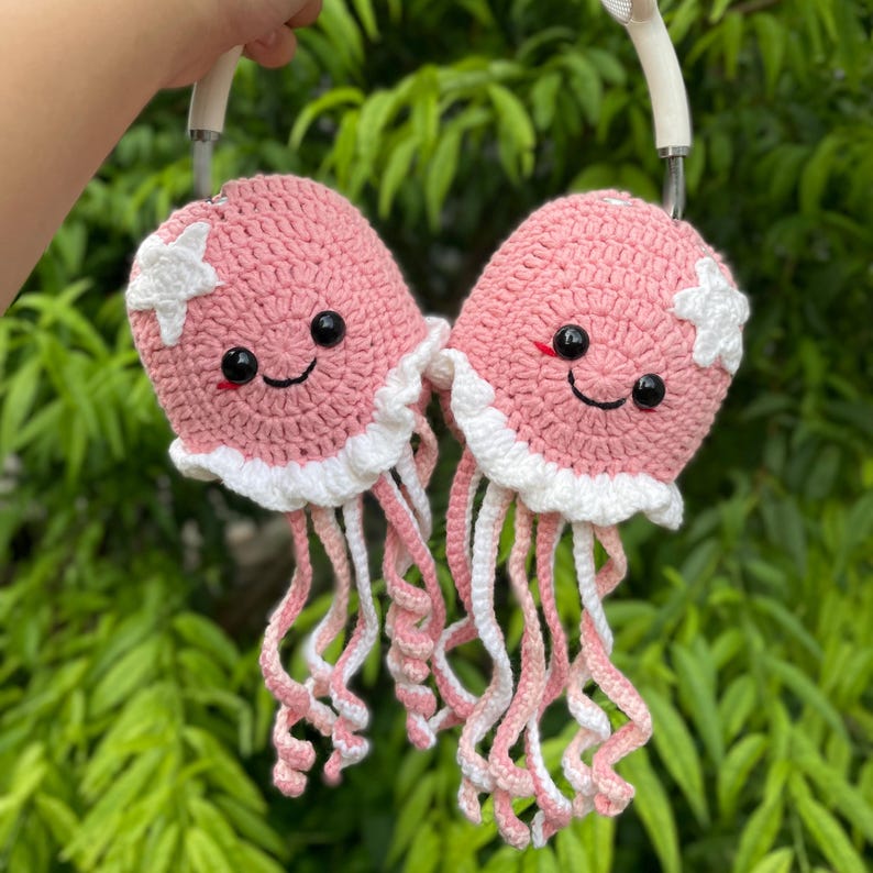 Puede incluir: Dos fundas de auriculares de ganchillo rosa con forma de medusas sonrientes. Cada funda tiene volantes blancos, una estrella blanca y tent&aacute;culos rosas y blancos colgantes. Los auriculares est&aacute;n sostenidos por una mano.