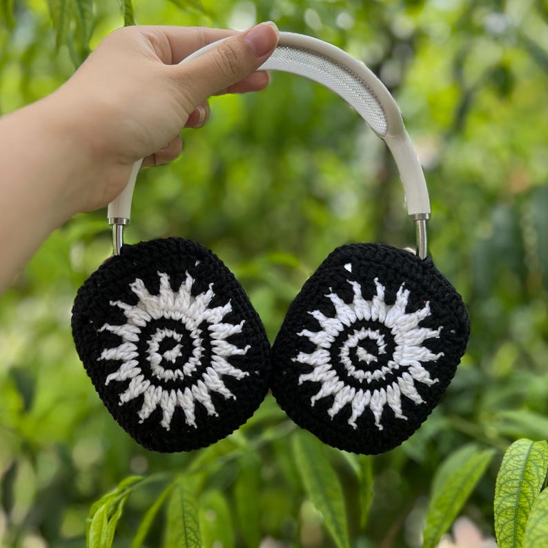 May include: Black and white crochet headphone covers with a sunburst design. The headphones have a white headband and silver accents. The covers are held up against a background of green foliage.