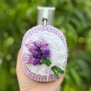 May include: A crocheted lavender-themed item featuring a bouquet of purple flowers with green stems tied with a light purple ribbon. The item has a white and light purple background and a crocheted border.