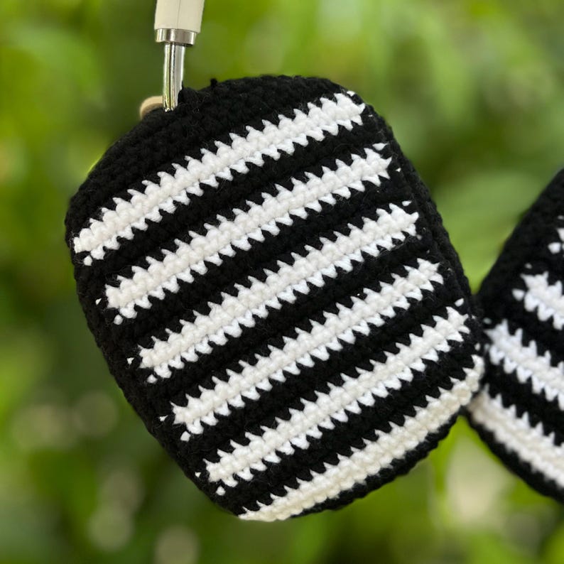 May include: Black and white striped crochet headphone case. The case is made of black and white yarn, with a repeating striped pattern. The case is hanging from a silver metal hook.