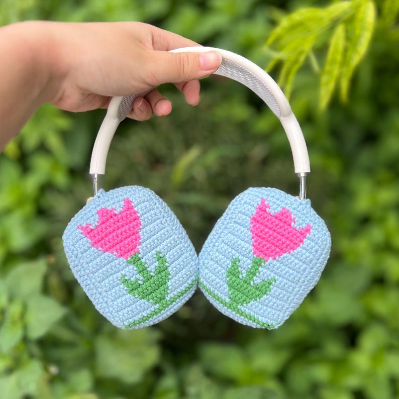May include: A pair of white headphones with light blue crochet ear covers. Each cover features a pink tulip with green leaves. The headphones are held against a blurred green background.