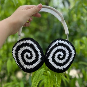 May include: White and silver headphones with black and white crocheted ear covers. The ear covers feature a spiral design. The headphones are held against a green, leafy background.