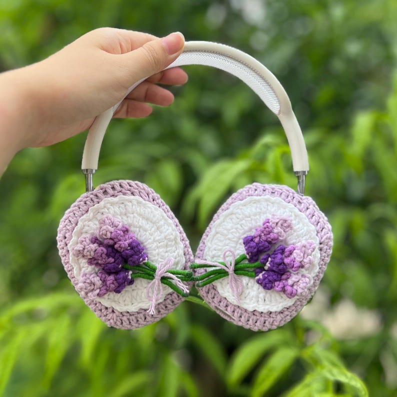 May include: White headphones with crochet ear covers. The covers feature a white center with a lavender and purple floral design. The crochet work is detailed, with a light purple border and green stems.