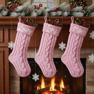 Pode incluir: Três meias de Natal rosa de malha de cabo penduradas acima de uma lareira. Cada meia tem uma punho branco com um nome bordado: "Lucy", "Ivy" e "Mom". As meias são decoradas com azevinho e bagas.