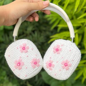 Puede incluir: Auriculares blancos con un diseño de ganchillo con flores rosas bordadas. Los auriculares tienen una diadema y auriculares blancos, con un patrón floral en los auriculares.