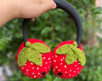 May include: A pair of red crocheted earmuffs shaped like strawberries, complete with green leaves and white seed details. The earmuffs have a black headband.
