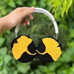 Pu&ograve; includere: Cuffie all'uncinetto nere e gialle con fascia bianca. I padiglioni sono neri con un motivo a foglia di ginkgo gialla. Lo sfondo &egrave; un fogliame verde.