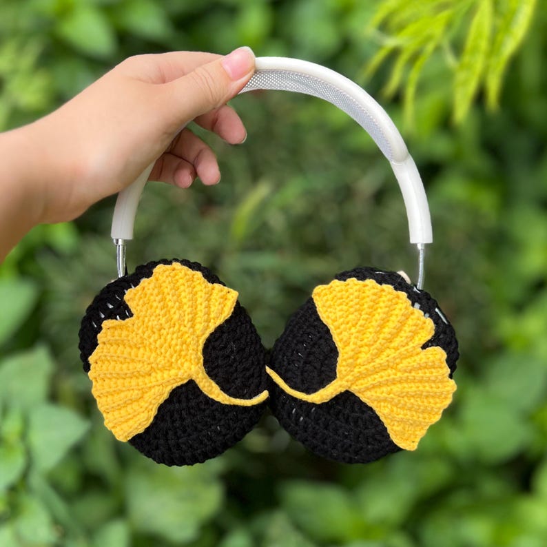 Pu&ograve; includere: Cuffie all'uncinetto nere e gialle con fascia bianca. I padiglioni sono neri con un motivo a foglia di ginkgo gialla. Lo sfondo &egrave; un fogliame verde.