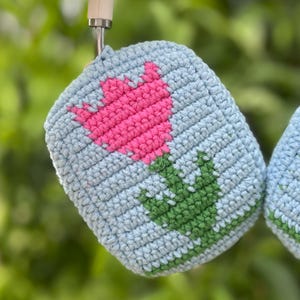 May include: A light blue crocheted pot holder with a pink tulip design and green leaves. The pot holder is made of a textured yarn and is hanging from a metal hook. The design is a simple, yet charming floral pattern.