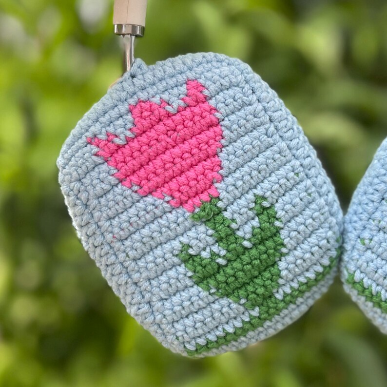 May include: A light blue crocheted pot holder with a pink tulip design and green leaves. The pot holder is made of a textured yarn and is hanging from a metal hook. The design is a simple, yet charming floral pattern.