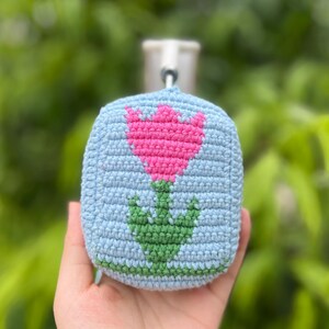 May include: A crocheted, light blue square with a pink tulip design and green stem. The handmade item has a small metal loop at the top for hanging. The background is a blurred green.