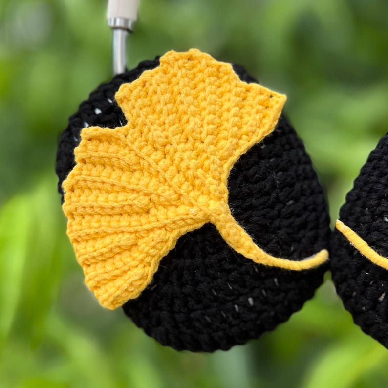 Pu&ograve; includere: Cuffie nere all'uncinetto con un design a foglia di ginkgo gialla. La foglia &egrave; finemente lavorata all'uncinetto e risalta sullo sfondo scuro. Le cuffie sono sospese su uno sfondo verde sfocato.