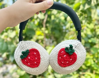 Könnte beinhalten: Gehäkelte Ohrenschützer mit einem schwarzen Stirnband und Ohrenschützern. Jeder Ohrenschützer hat eine rote Erdbeere mit grünen Blättern. Die Ohrenschützer werden vor einem Hintergrund aus grünem Laub gehalten.