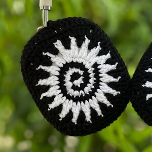 May include: Black and white crochet headphone covers. The covers feature a sunburst design with a black background and white detailing. The headphones are hanging against a blurred green background.