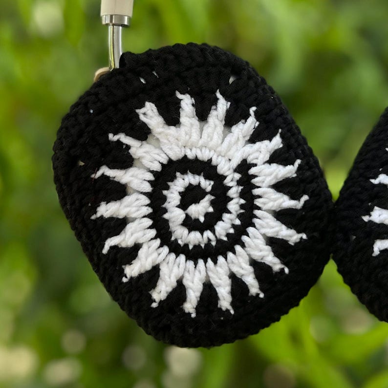 May include: Black and white crochet headphone covers. The covers feature a sunburst design with a black background and white detailing. The headphones are hanging against a blurred green background.