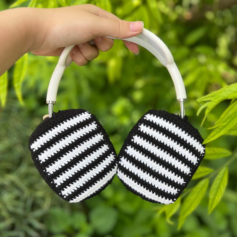 May include: Headphones with black and white striped ear covers. The headphones have a white headband and silver accents. The ear covers are crocheted with a black background and white stripes. The headphones are held up by a hand in front of a green leafy background.