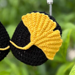 Pu&ograve; includere: Cuffie nere all'uncinetto con un design a foglia di ginkgo giallo brillante. La foglia &egrave; realizzata all'uncinetto in modo intricato, aggiungendo un tocco unico e fatto a mano. Le cuffie sono ambientate su uno sfondo verde sfocato, evidenziando i colori e le trame contrastanti.